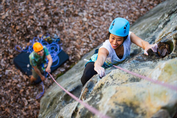 A young asian woman in a blue helmet climbs a rock cliff on top rope while her partner wearing an orange helmet belays from below, seen from above
