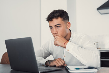 Thoughtful businessman touching chin, pondering ideas or strategy, sitting at wooden work desk with laptop, freelancer working on online project, student preparing for exam at home
