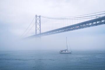 View of 25 de Abril Bridge famous tourist landmark of Lisbon connecting Lisboa and Almada in heavy fog mist wtih yacht boats passing under. Lisbon, Portugal