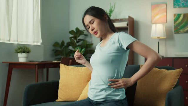 Young Asian woman hand holding her low back, feeling low back pain and hurt while sits on sofa in living room at home. Asian female suffering, relief  from back pain. Health care and medical concept.