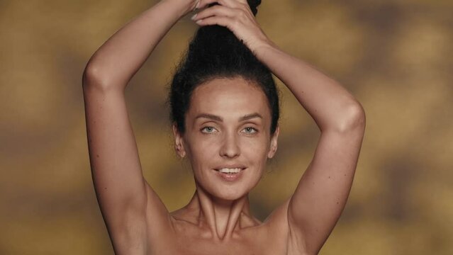 A brunette woman with a bun on her head, unbind her thick long curly hair. Portrait of a smiling seminude woman in the studio on a yellow background with highlights. Slow motion.