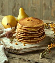 Beautifully presented stack of oatmeal pancakes on a light-colored wooden board. A sprig of sea buckthorn and a honey spoon are put next to it. Recipe Blog Post. Image for food selling company.