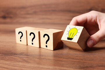 Idea concept. Woman putting cube with illustration of light bulb to other ones with question marks on wooden table, closeup