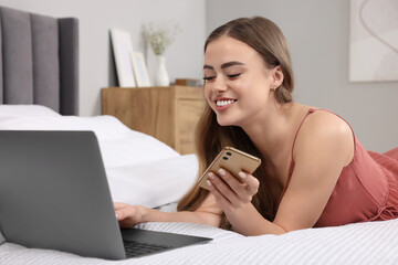Happy woman with smartphone and laptop on bed in bedroom