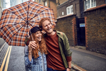 Young caucasian couple hiding under an umbrella while walking on a city street during rain
