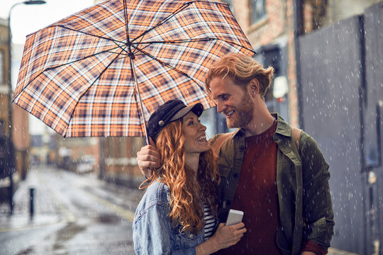 Young caucasian couple hiding under an umbrella while walking on a city street during rain