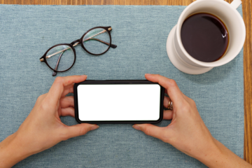 A woman's hands hold a mobile smartphone with transparent screen while her glasses and a cup of coffee rest gently on the table before her, accompanied by serene scene