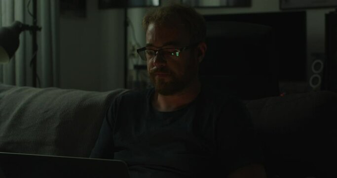 A man works on his laptop in a very dark living room, he turns his head towards a noise