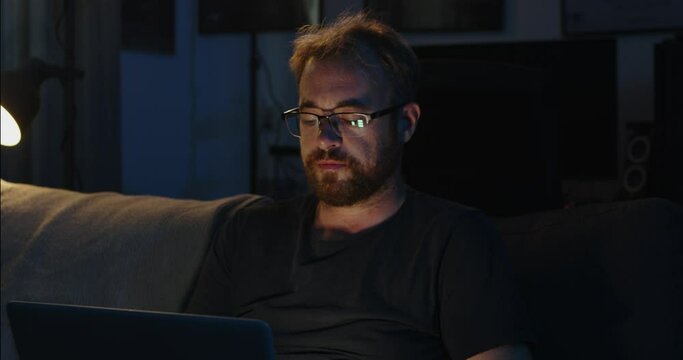 A man is sitting in his living room couch and working on his laptop late at night. He turns his head towards something
