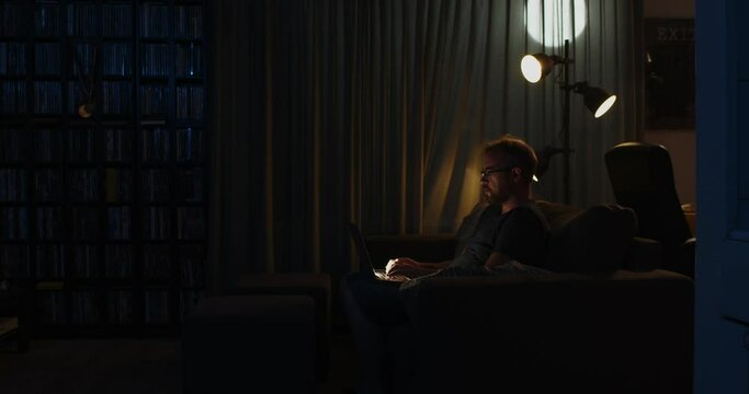 Workaholic man working on a laptop in his living room late at night. A bright moon shines through the curtains while the camera slowly zooms out