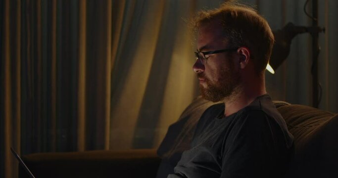 Close up of a man working on his laptop on his couch late and night. He slowly looks up