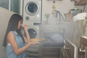 Fototapeta na wymiar Housewife failed to cook : Young Asian housewife in the kitchen baking bread in the microwave oven the appliance set the baking time too long until the bread burns there is smoke in the kitchen.