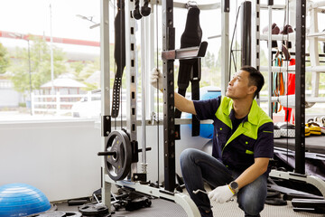 Professional Asian male service worker or fitter checks equipment, maintains and secures fitness equipment in indoor gym, provides safety for users : Skilled technicians repair exercise machines. - Powered by Adobe