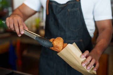 Close-up of Indian man hands preparing bread and croissants in paper bags. to prepare for delivery to customers ordering online behind the bar counter in a coffee shop small family business