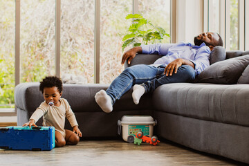 Tired African-American father naps on the couch because of the necessity of raising his naughty...