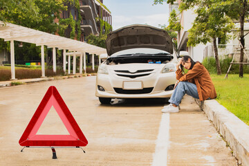 Asian woman open hood car parked road and sits stressed in front parked car breakdown without knowing the cause during the journey by placing warning triangles prevent danger from oncoming vehicles.