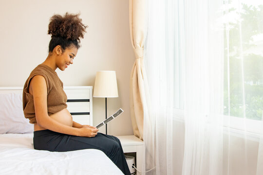 Side view happy portrait African American mother pregnant sits in bed looking at ultrasound of her upcoming baby with anticipation and delight to see the evolution of a healthy fetus.