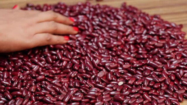 A woman holds a pile of red beans on a wooden table, while a hand touches another pile nearby
