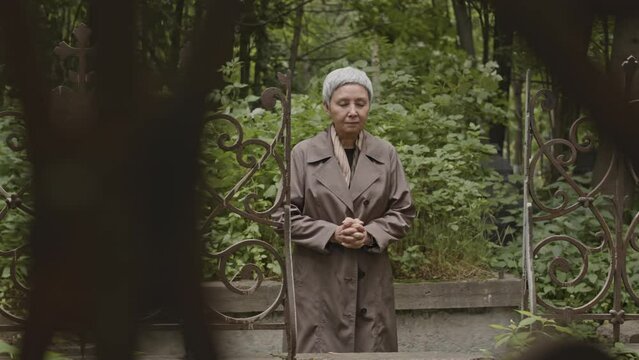 Medium shot of mourning senior Asian woman standing by metal gates at cemetery with locked hands