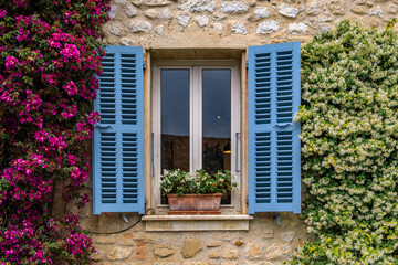 Fototapeta na wymiar Bougainvilia and jasmine flower vines framing an old stone house window in the medieval town of Saint Paul de Vence, French Riviera, South of France