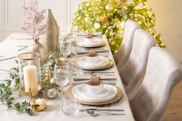 Table set for Christmas dinner, dinnerware set on gold charger plates in white and gold colors with candles, inside a Scandinavian house interior - Powered by Adobe