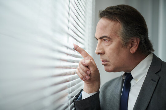 Curious mature businessman peaking through window blinds - Powered by Adobe