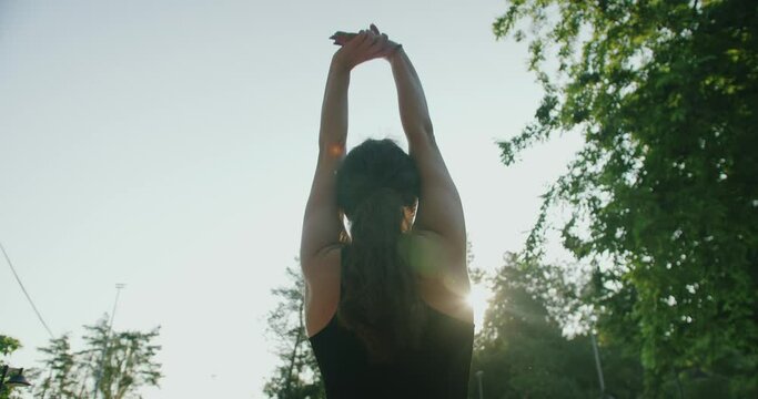 Back view on sporty woman brunette exercising outdoors, warm up and stretching arms. Young female have fitness training at nature in sunny morning. Healthy lifestyle concept