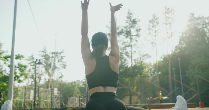 Back view on athletic woman in activewear doing leg split on exercise mat, raise hands up and stretching body outdoors in park. Young female exercising and training flexibility outside.