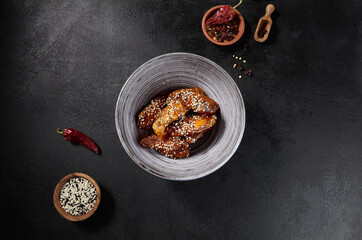 Horizontal top view of Buffalo wings glazed with honey and sprinkled with sesame seeds in a gray bowl on a black background with space for text