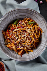 Overhead view in a vertical frame, capturing udon noodles with chicken and vegetables stir-fried in a garlic sauce. The black backdrop, chopsticks and cloth add depth and interest