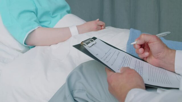 Cropped shot of medical worker writing symptoms of patient while filling health card sitting next to bed in ward