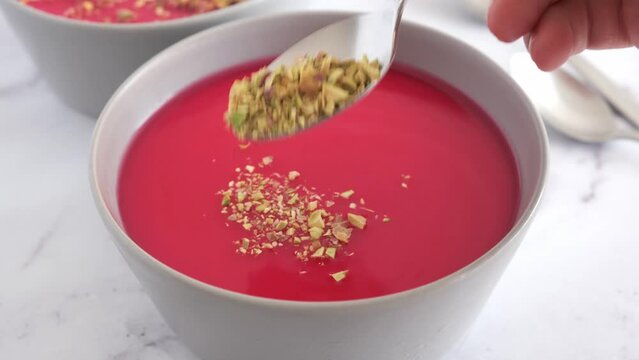 Cooking Malabi traditional Arabic dessert. Milk pudding with pink syrup and pistachio in gray bowl.