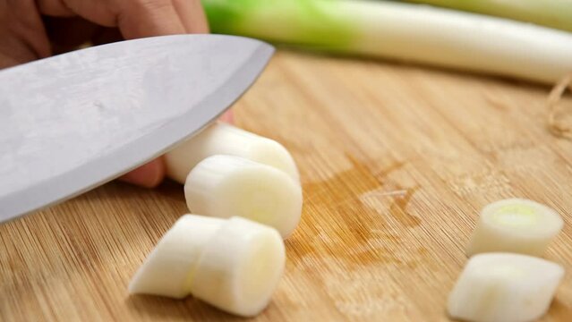 Woman's hand holding a knife cutting fresh Japanese scallions on a wooden chopping board. Sliced leek or Chopped green japanese bunching onion on a wooden board.
