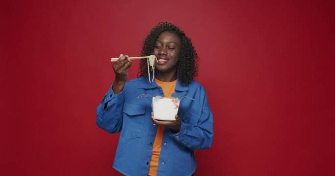 African american woman looking at camera and smiling, preparing to eat noodles, seafood and vegetables with chopsticks on isolated red background. Young woman eating Asian noodles for lunch.