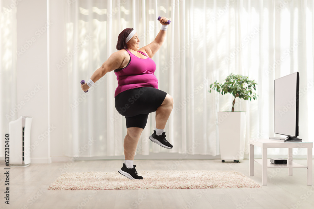 Poster cheerful overweight woman exercising with small dumbbells in front of tv