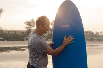 Middle aged athlete standing on sandy beach at sunset and touching blue surfboard, checking...