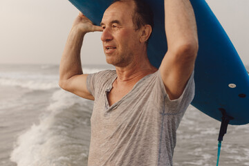Portrait of mature surfer looking ahead while carrying blue surfboard on head with hands, standing...