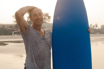 Happy mature man with surfboard standing on sandy beach and touching bald head while looking at...