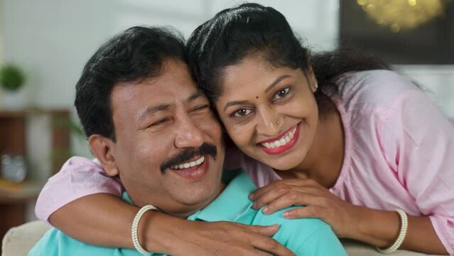 Close up shot of happy indian middle aged couple embracing or hugging by looking at camera at home - concept of family bonding, togetherness and relaxation