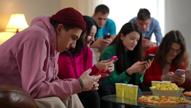 Side view portrait of concentrated young man with blurred focused friends surfing Internet in phone apps. Young Caucasian men and women scrolling social media partying indoors