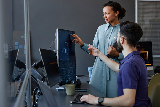 Smiling computer programmer gesturing and explaining to colleague over desktop PC