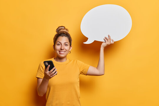 Waist up shot of good looking young European woman with hair bun holds mobile phone and blank communication bubble for your advertising content wears casual t shirt isolated over yellow background.
