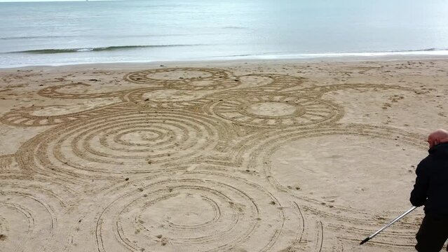 Aerial view circling man creating interesting zen sand art design on windy Welsh golden beach