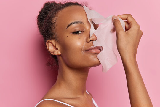 Beautiful woman with curly combed hair puts on facial mask for skin treatment smiles gently wears t shirt isolated over pink wall. Young female model removing from face skincare hydrating sheet mask