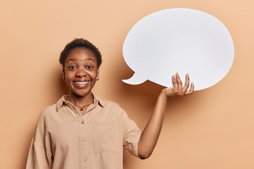 People emotions concept. Portrait of young happy smiling African american woman wearing blouse standing isolated on beige background holding white speech bubble with blank space for your promotion
