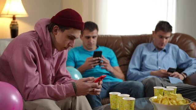 Side view focused young Caucasian man absorbed with smartphone app with blurred friends sitting at background surfing Internet. Careless mates on weekend ignoring each other