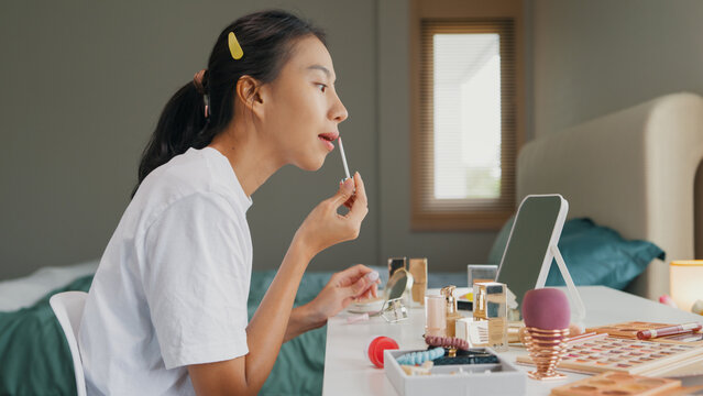 Young happy Asia girl beauty blogger, look the mirror, makeup apply lipstick balm on her lips in bedroom. Lifestyle and people, Make up Natural look, Skin Care Cosmetics, Morning routine concept.