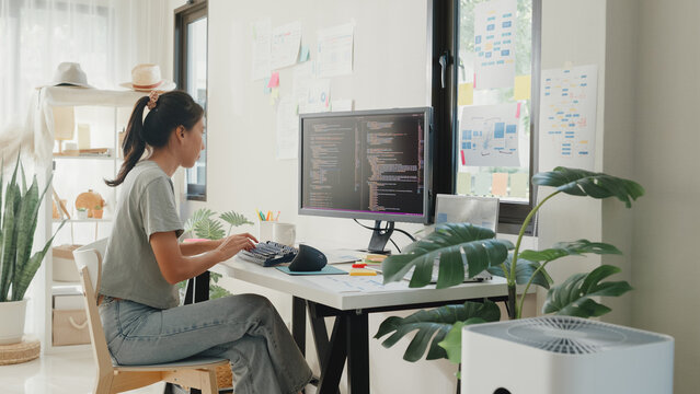 Side view of professional young Asia girl IT development programmer typing on keyboard coding programming fixing data code on computer screen and laptop on table in workroom at house office.