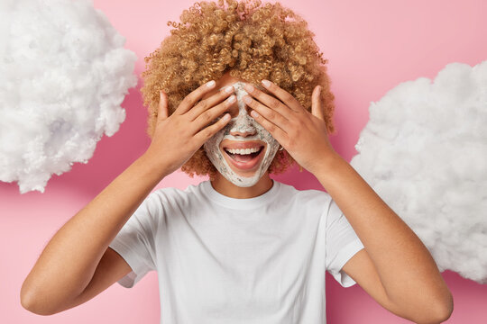 Beauty and cosmetology procedure. Indoor photo of young happy smiling African american girl applying clay face mask closing eyes with hands standing in centre isolated on pink background with clouds