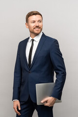 Smiling man in business suit with laptop on studio background. Confident male businessman successful entrepreneur look away on empty space with tablet in hand standing isolated grey wall blank